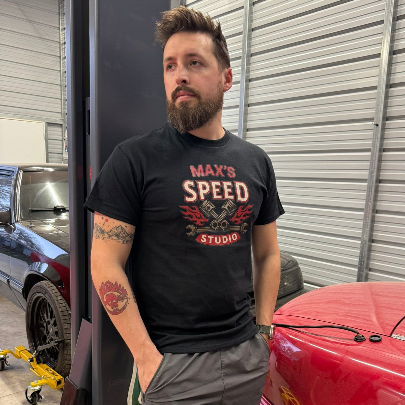 Man wearing a black t-shirt with 'Max's Speed Studio' in a garage with red car and equipment.