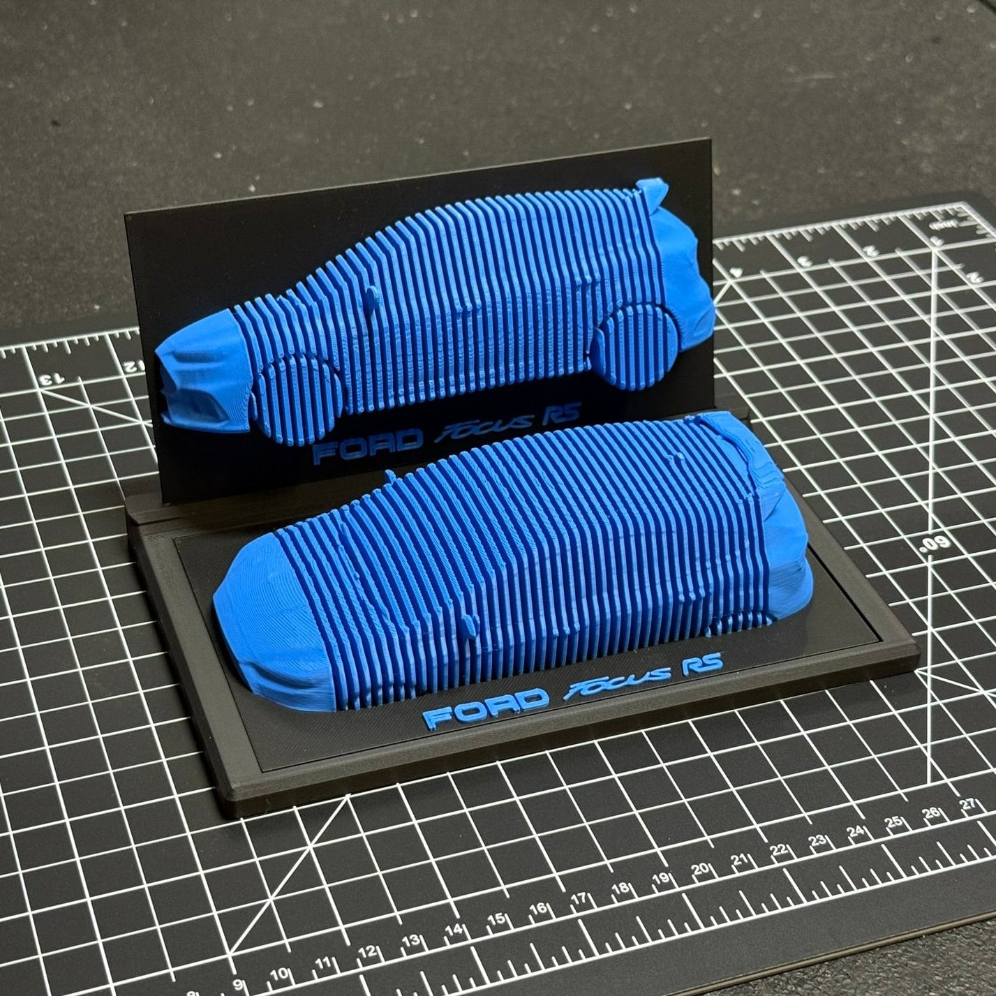 Blue 3D-printed car model on a black display card with 'Ford Focus RS' branding, placed on a grid surface.