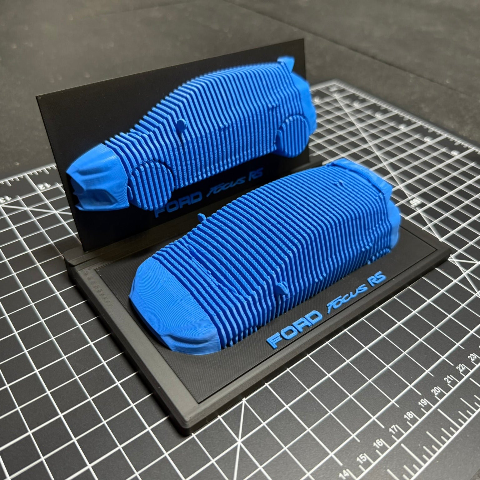 Blue 3D-printed car model on a black stand with 'Ford' branding, placed on a grid surface.